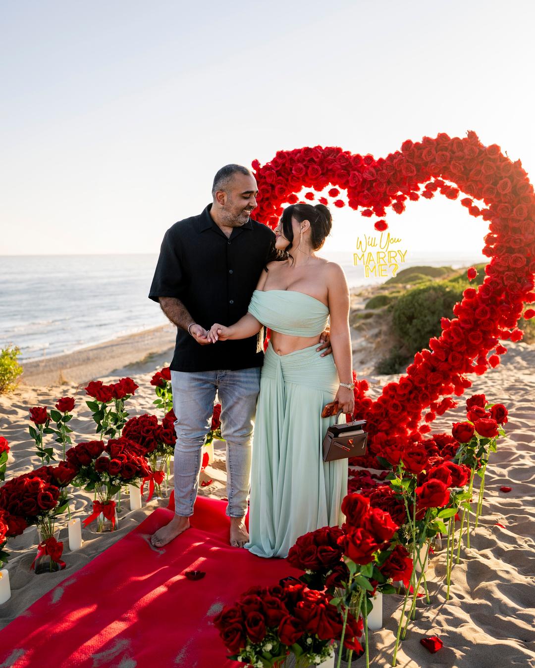 Newly engaged couple on a red carpet in front of a heart of red roses on the beach