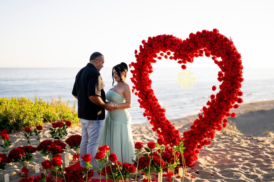 Couple holding hands in front of a heart of red roses on the beach, the sea behind them