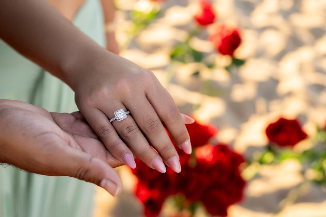 Close-up of a diamond ring on her hand, red roses in the sand behind