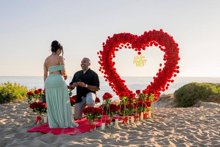 He kneels with the ring beside a heart of red roses as she turns towards him